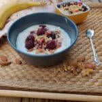 יוגורט סויה בתוך קערה עם פירות אדומים וגרנולה מעל כפית מתכת בצד, מונח על לפייסמנט דמוי עץ