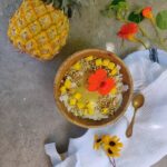 סמודי בול אננס בצבע צהוב מוגש עם פירות מעל וחתיכות אננס. מוגש בתוך קערה דמוי עץ וכפית זהב ליד על מפה לבנה ושיש אפור
