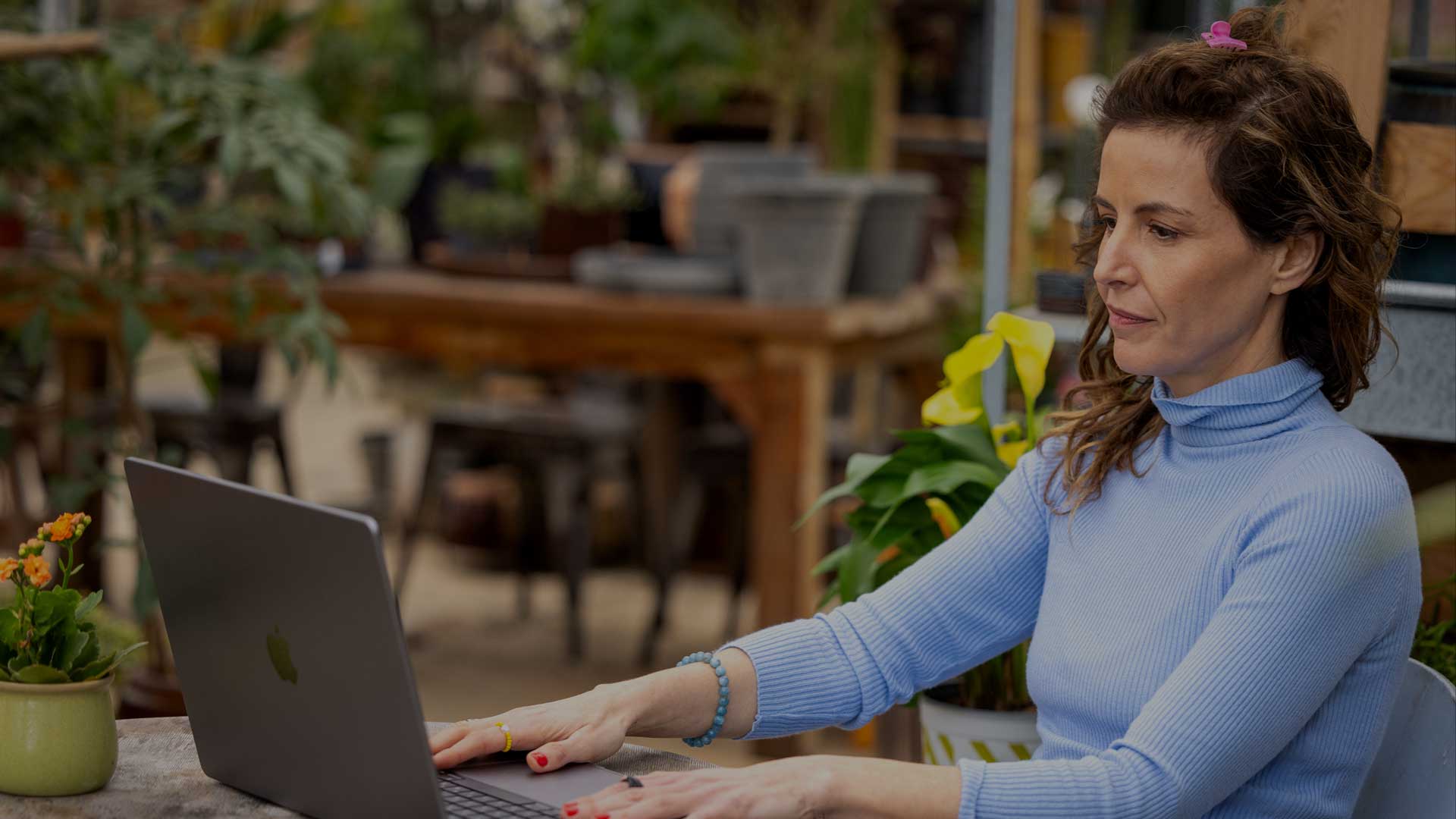 ליבי לוי נטורופתית יושבת בחצר עם המחשב על שולחן עץ לבושה חולצה כחולה ארוכה ועצים ברקע