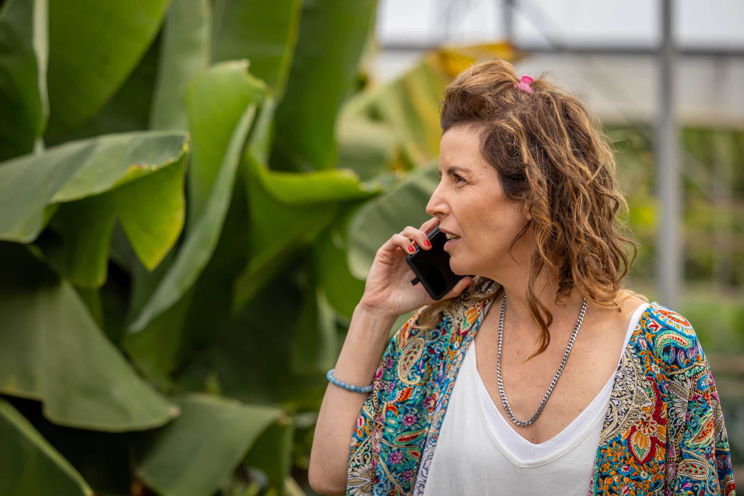 ליבי לוי נטורופתית מדברת בפלאפון בטבע ליד עלים ירוקים גדולים, לבושה גופיה לבנה ועליונית פרחונית ירקרקה