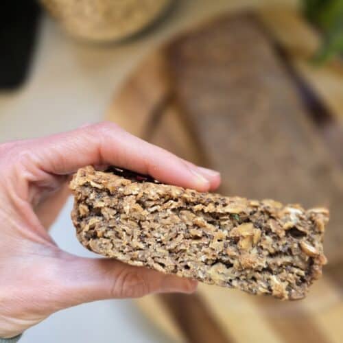 דייסה אפויה בצורת עוגה, עם זרעי צ׳יה, פרוסה מוחזקת בין שתי אצבעות וברקע שאר העוגה מונחת על צלחת עץ